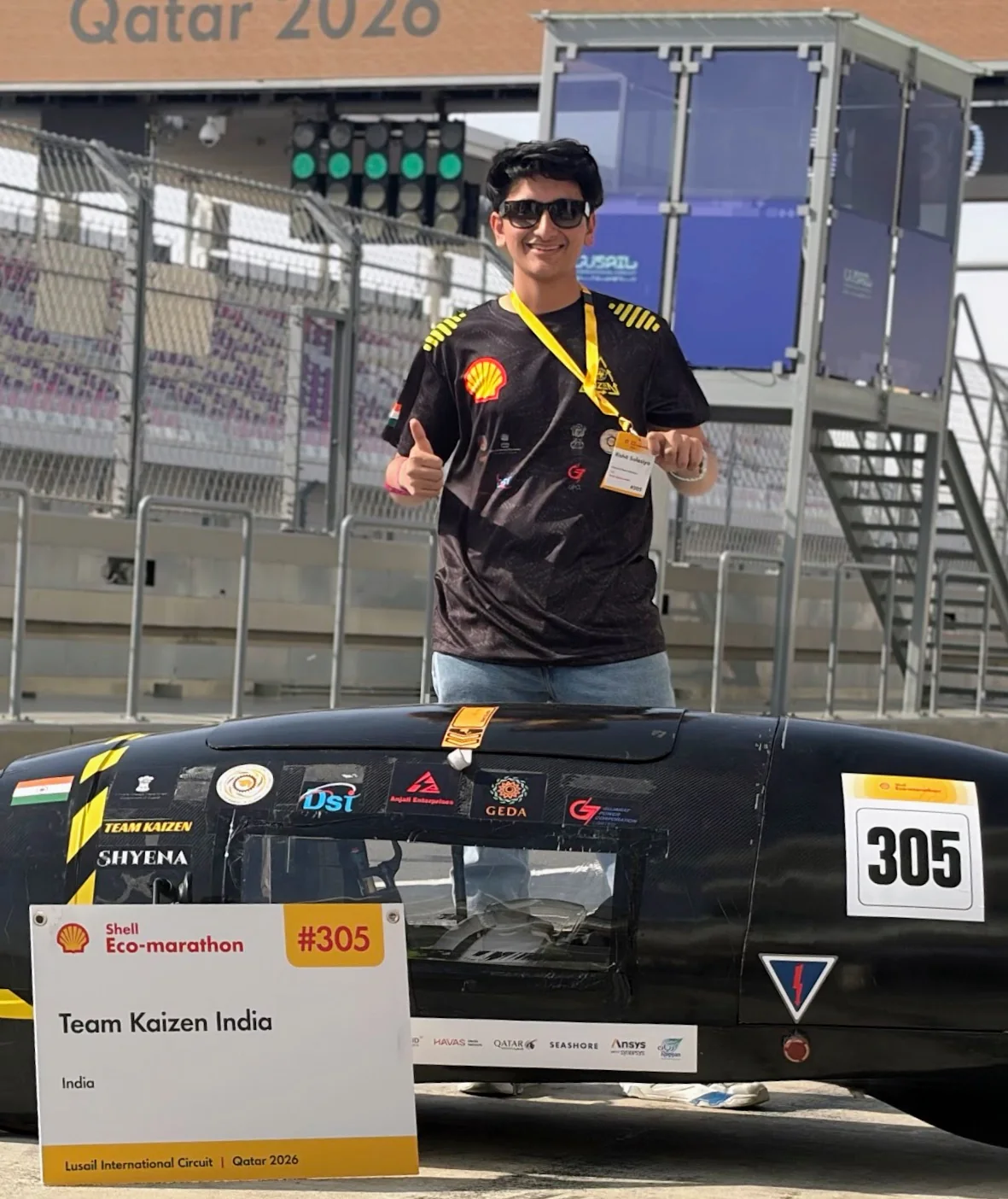 Rishit Salasiya giving a thumbs-up behind SHYENA, Team Kaizen India's car #305, in the pit lane at Shell Eco-marathon Qatar 2026, Lusail International Circuit